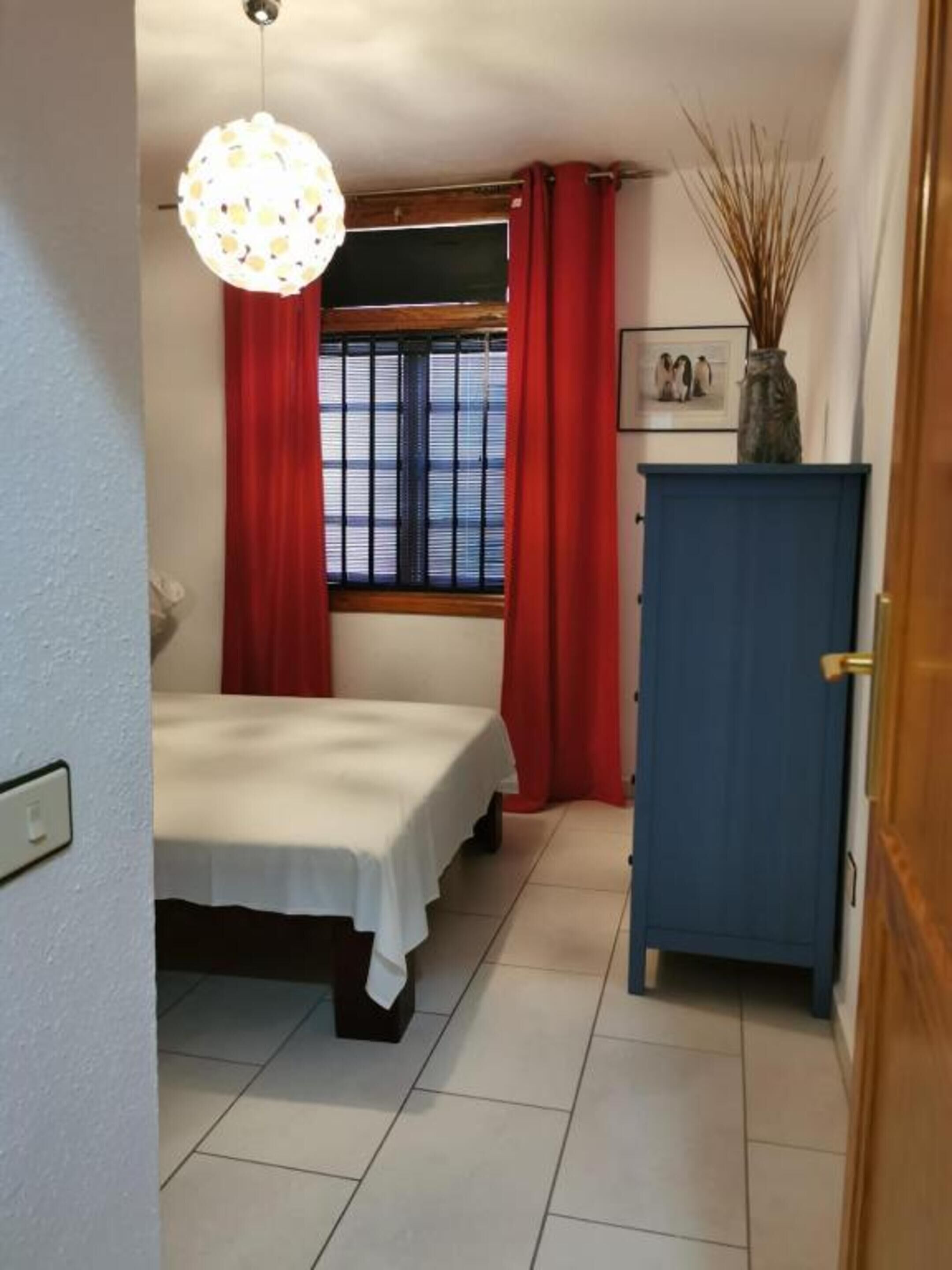 A bedroom with a bed, a blue cabinet, a window with red curtains, and a hanging light fixture.