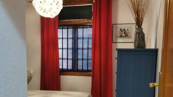 A bedroom with a bed, a blue cabinet, a window with red curtains, and a hanging light fixture.