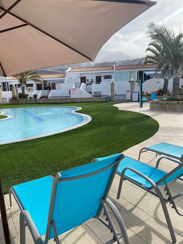 A pool area with artificial grass, a covered seating area, and a view of a residential building in the background. - Apartamentos Malibu Park (Adeje)