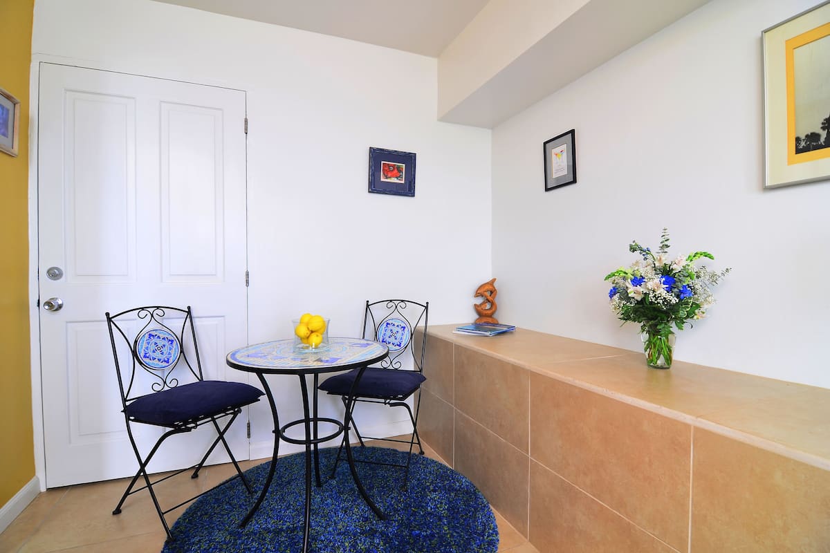 A small room with a white door, two chairs, a small table, a blue rug, and a vase of flowers on a tiled counter.