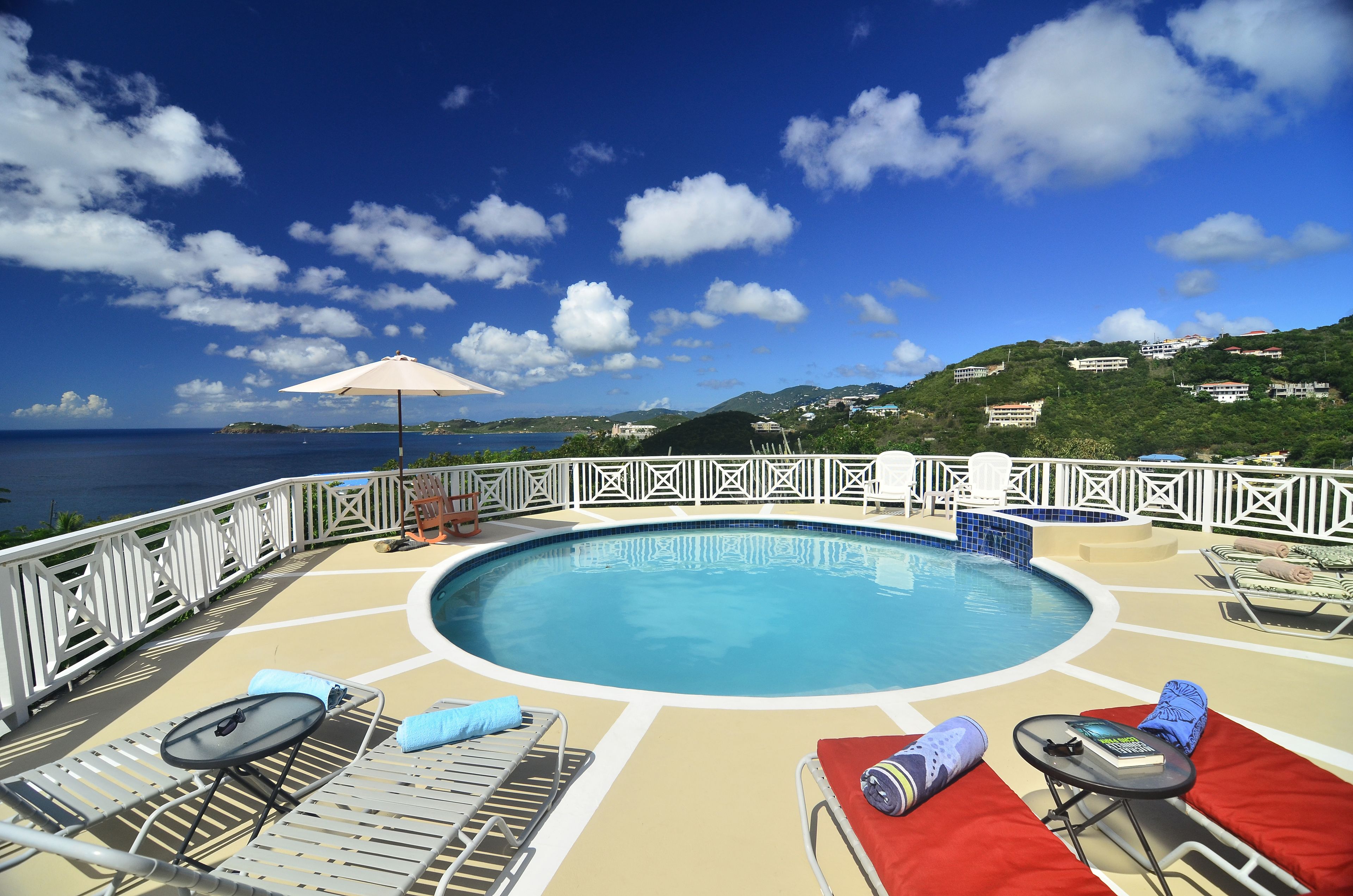 A pool area with lounge chairs, a small table, and a view of the ocean and hills.