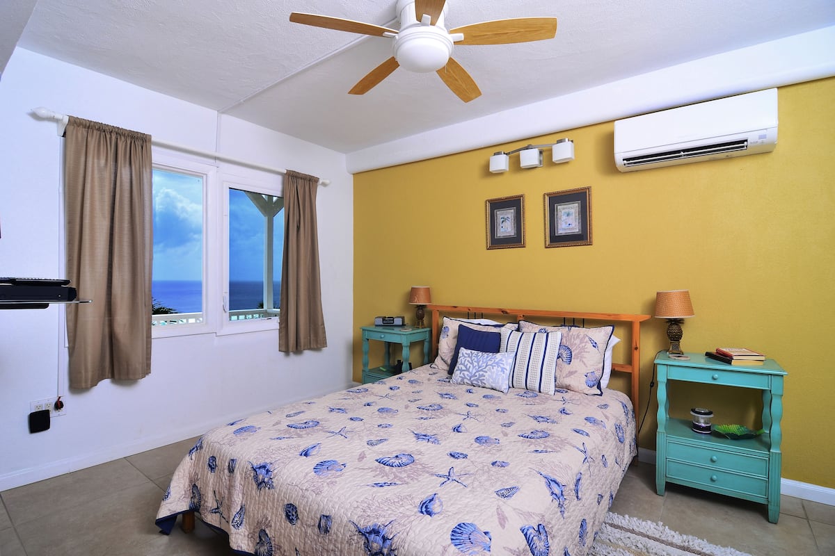 A bedroom with a bed, a ceiling fan, an air conditioner, a nightstand, and a window with curtains.