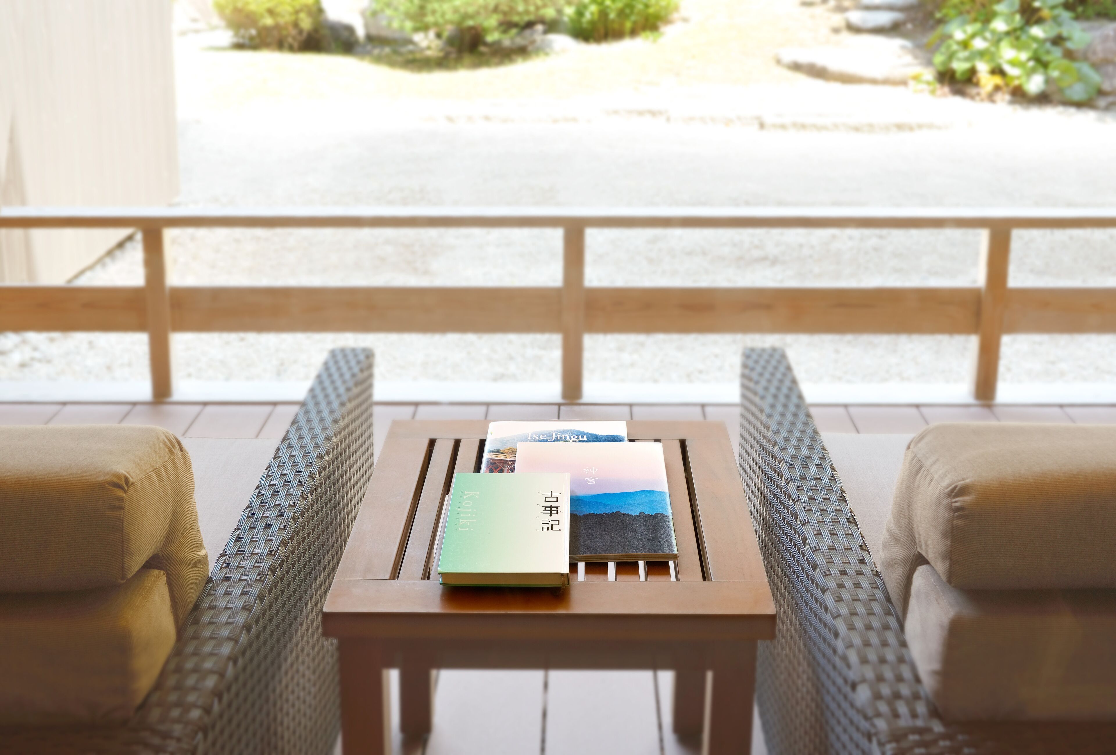 A wooden table with three books and two cushioned chairs in a room with a view of a garden.
