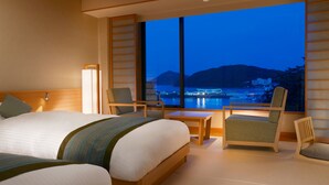 Habitación de hotel con una cama grande, dos sillones, una mesita y un ventanal con vistas a un paisaje urbano y montañas al anochecer.