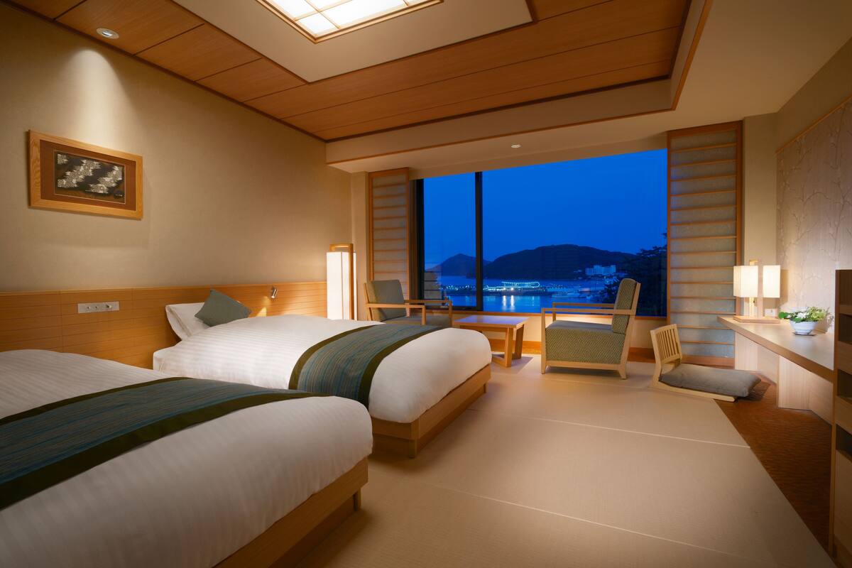 A hotel room with two beds, a desk, a chair, a nightstand, and a large window with a view of a body of water and hills.