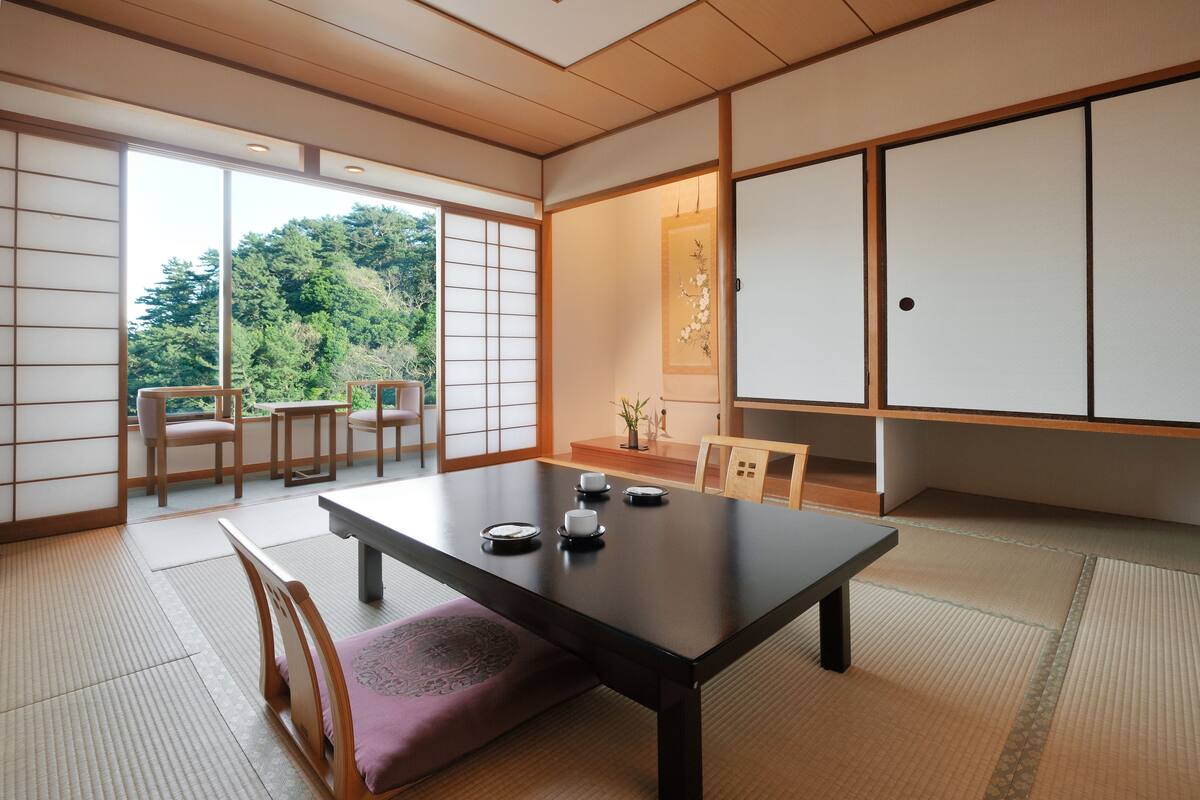 A traditional Japanese room with tatami flooring, a low table with tea set, sliding doors, and a view of greenery outside.