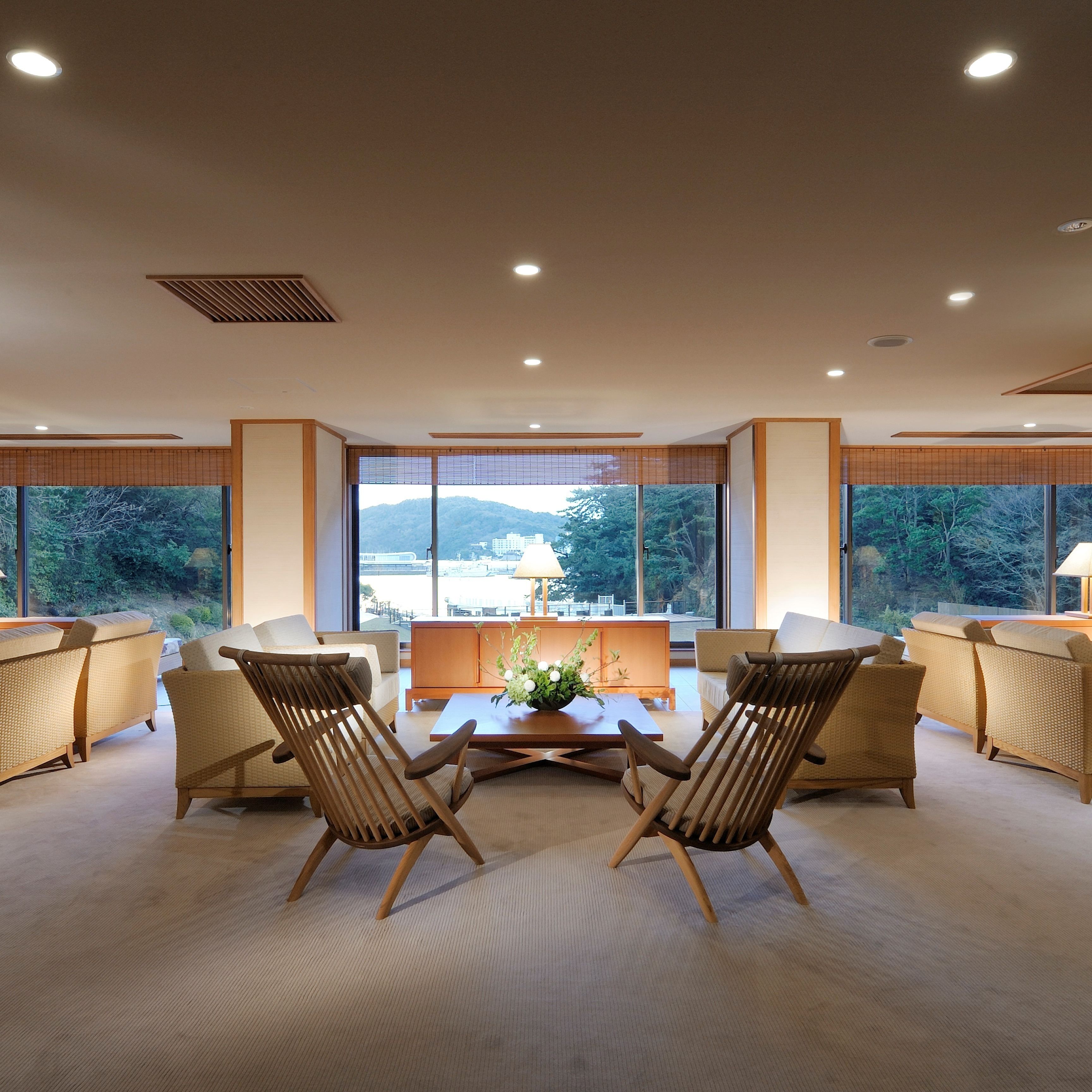 A modern living room with a view of a mountainous landscape through large windows, featuring a central coffee table, two wooden chairs, and a sofa.