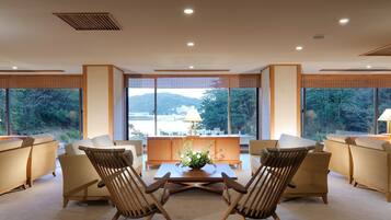 A modern living room with a view of a mountainous landscape through large windows, featuring a central coffee table, two wooden chairs, and a sofa.