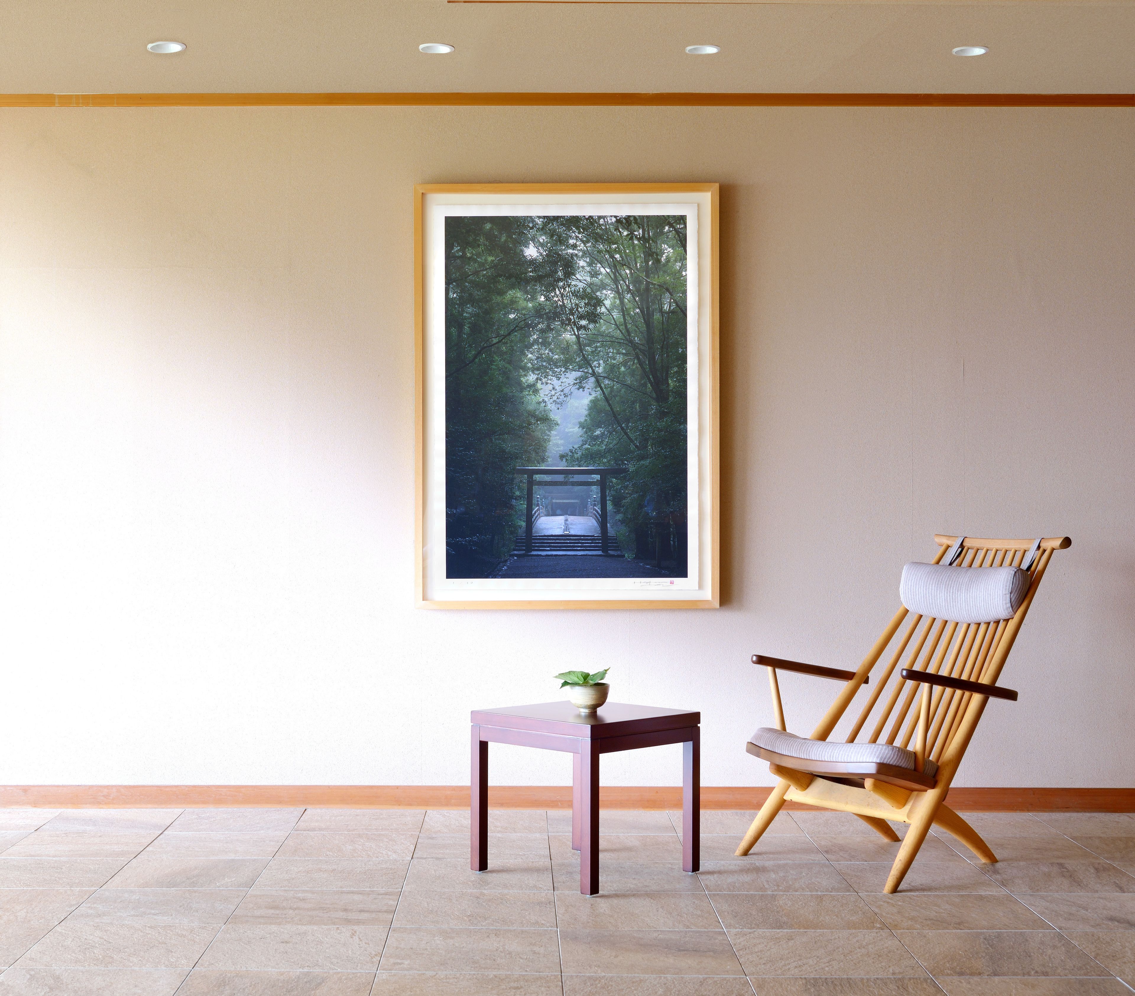 A room with a framed picture of a forest pathway, a wooden chair with cushions, and a small table with a plant.