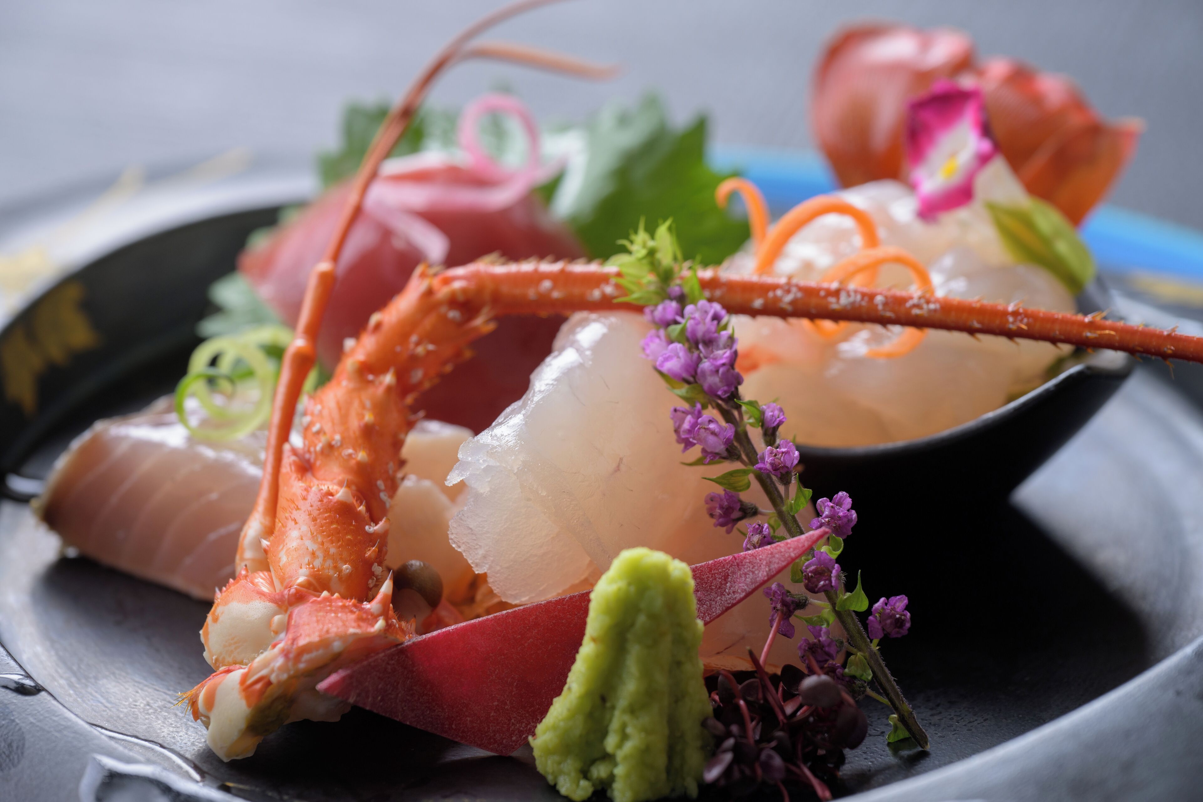 A seafood dish with a lobster tail, sashimi, and garnishes on a black plate.