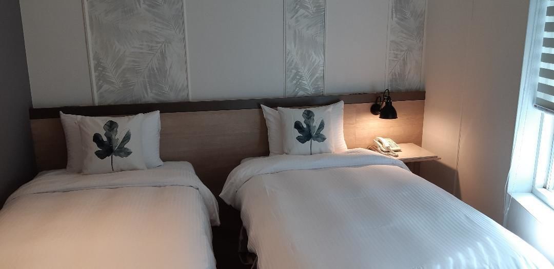 Une chambre avec un grand lit double, composé de deux lits individuels, chacun agrémenté d’un oreiller orné d’un motif de feuille. On y trouve une table de chevet avec une lampe et un téléphone.
