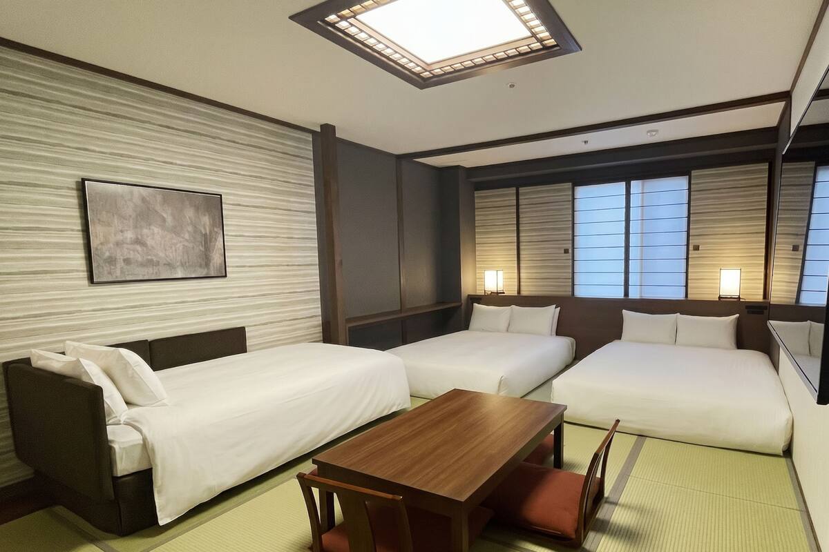 A hotel room with two beds, a wooden table, tatami mats, a wall-mounted artwork, and a ceiling light.