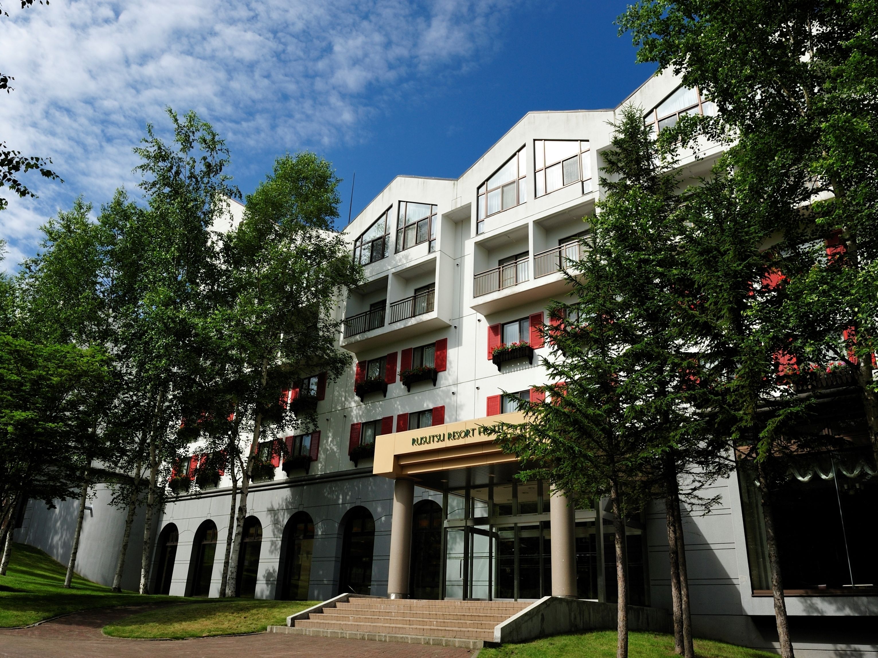 A multi-story hotel with a modern architectural design, featuring balconies with red shutters and a prominent entrance with a sign.