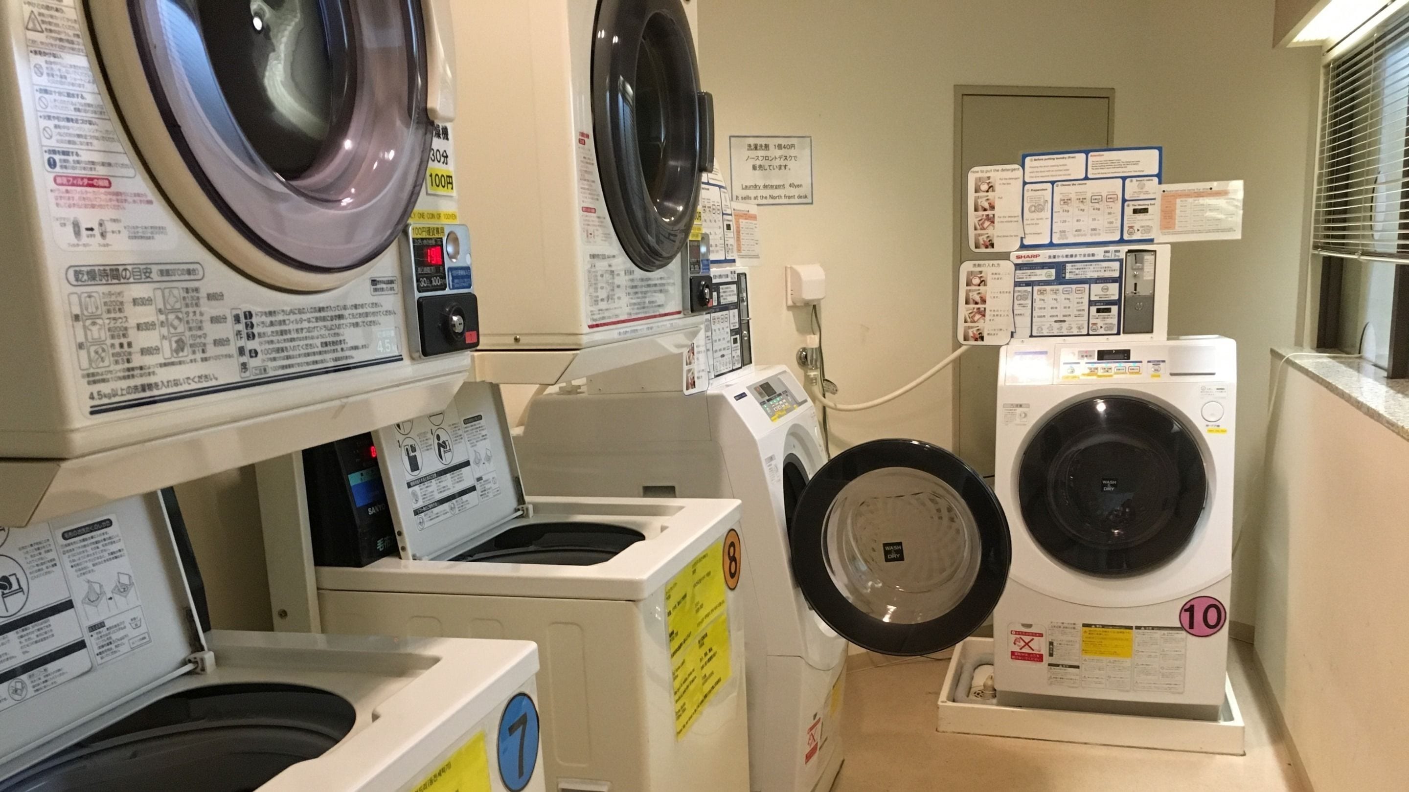 A laundry room with multiple washing machines, including a front-loading machine and a top-loading machine. There are instructional signs and a price tag visible.