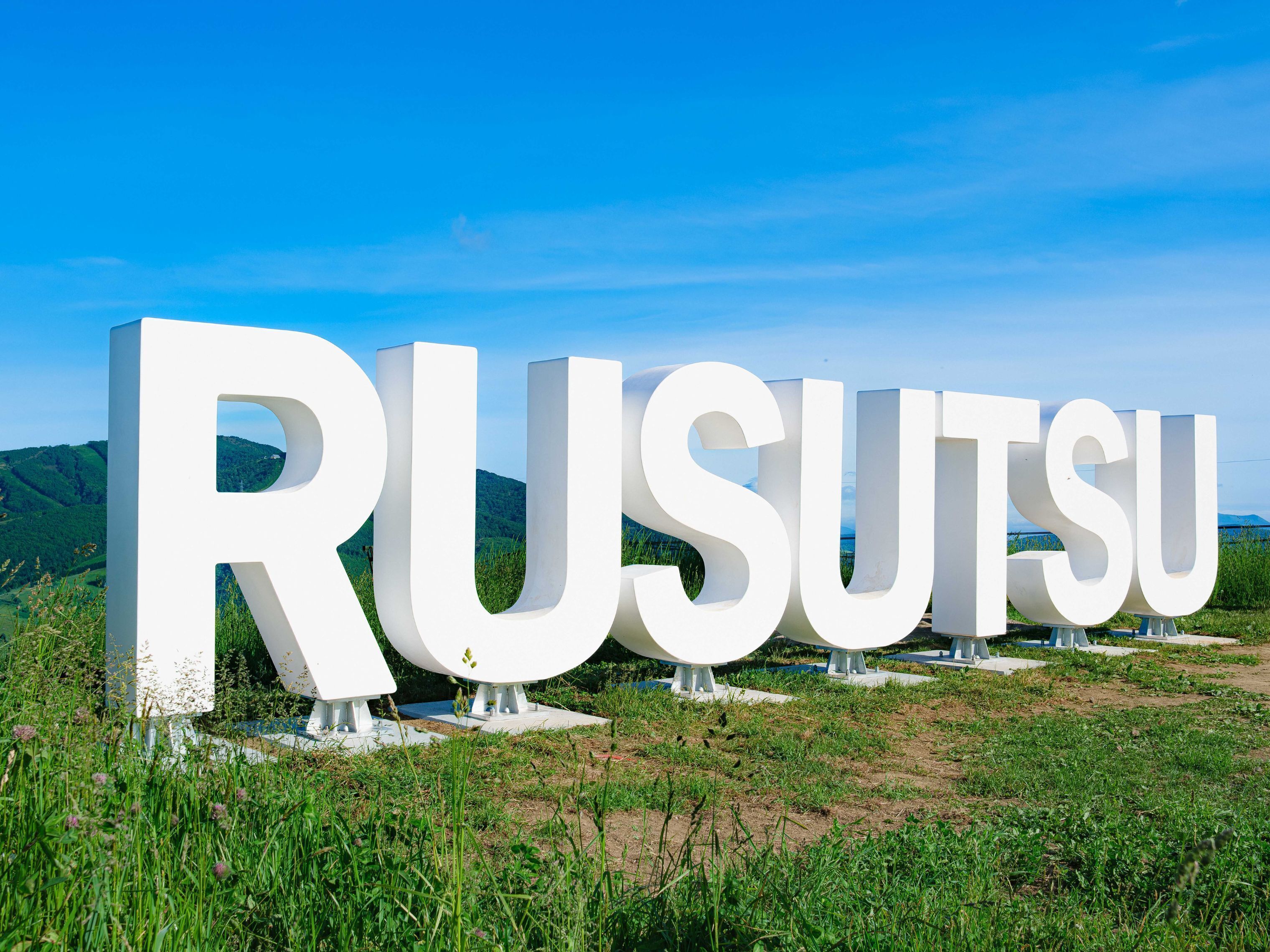 Large white letters spelling 'RUSUTSU' on a grassy field with a clear blue sky in the background.