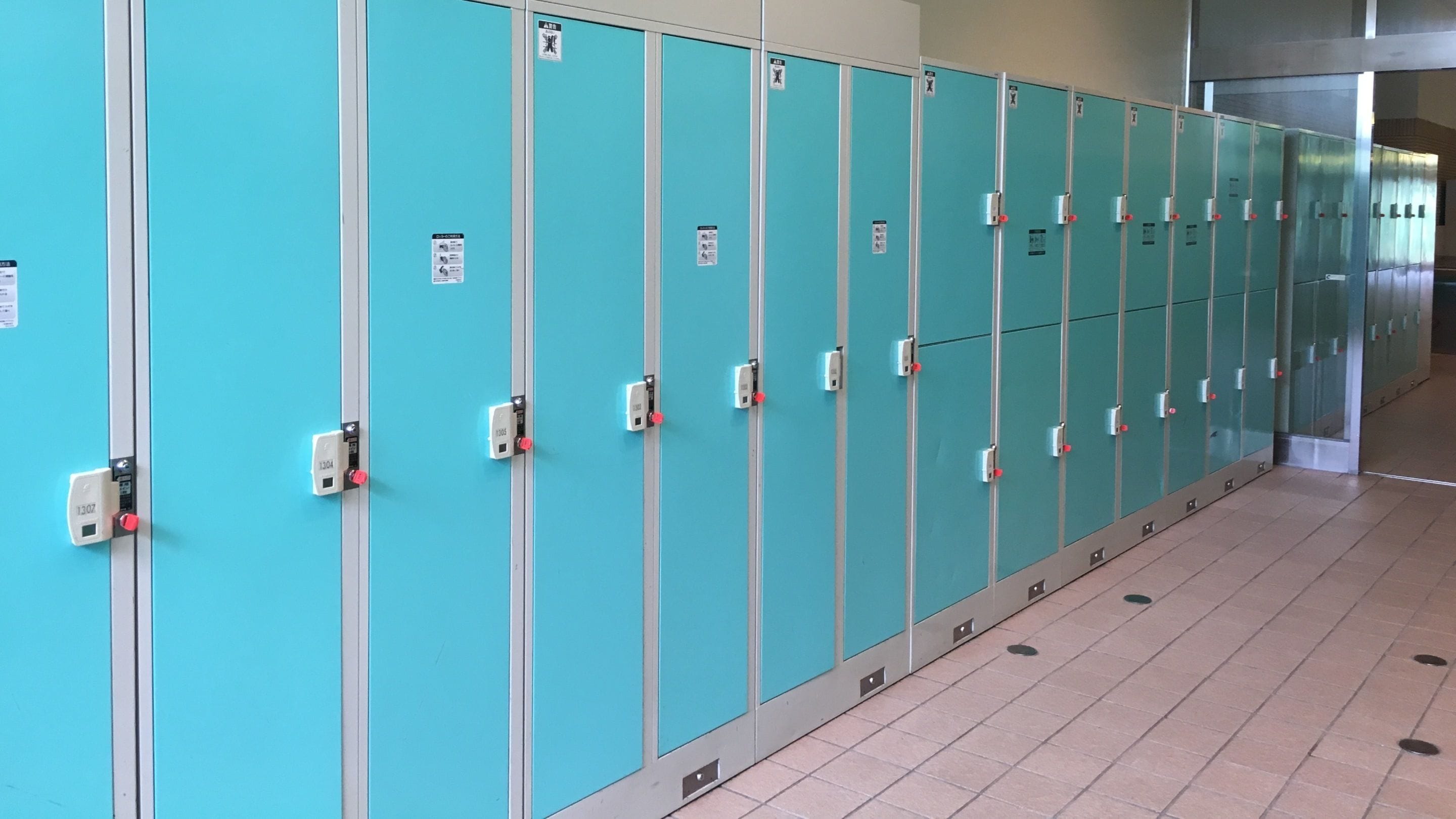A row of teal lockers with metal handles and numbered locks, lined up against a light blue wall in a hallway with tiled flooring.
