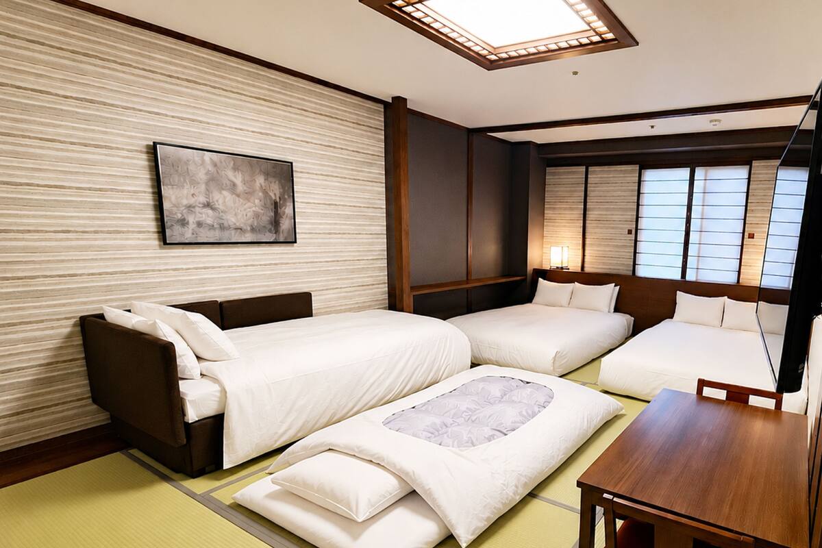 A hotel room with a traditional tatami mat floor, a sofa bed, a single bed, a wooden table, and a wall-mounted artwork.
