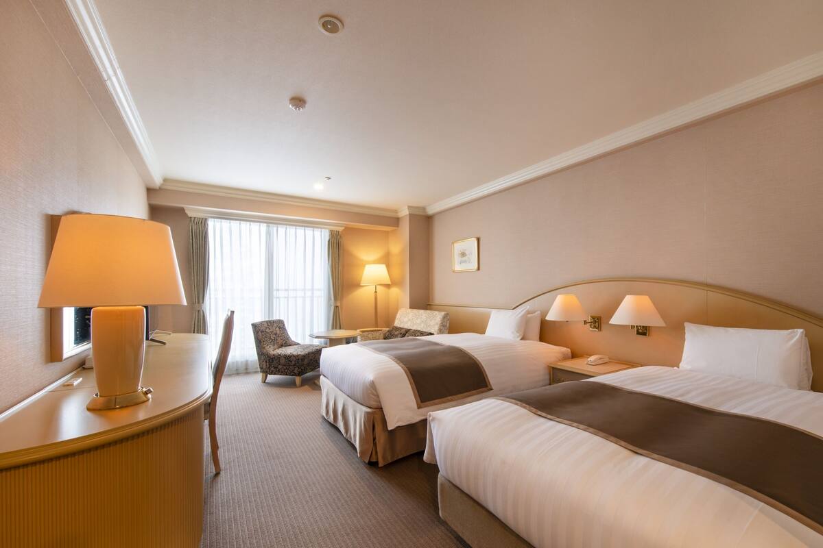 A hotel room with two beds, a desk with a lamp, a chair, a bedside table with a lamp, and a window with curtains.
