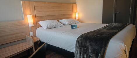 Ein Hotelzimmer mit einem hölzernen Kopfteil, zwei Nachttischlampen, einem Bett mit weißer Decke und grauer Tagesdecke, einem Nachttisch und einem Holzbüro.