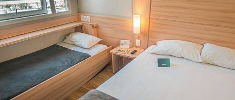 A hotel room with two beds, a bedside table with a lamp, and a window with a city view.