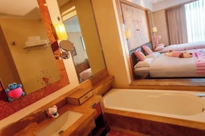 Une chambre d’hôtel avec un grand miroir, une baignoire, un lit avec une literie rose et blanche, et un petit bureau avec une lampe.