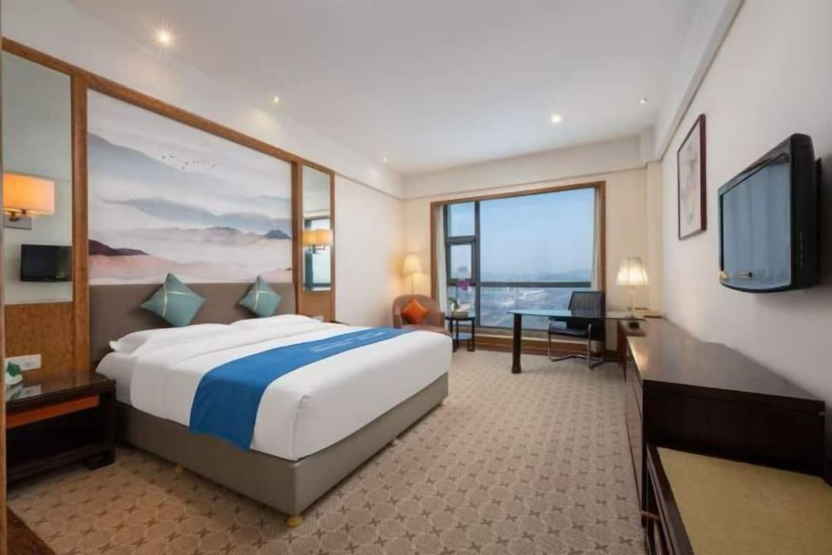 Een hotelkamer met een groot bed, een bureau, een stoel, een televisie en uitzicht op de stad door een groot raam.