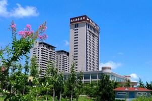 A multi-story hotel with a prominent building in the background, surrounded by greenery and flowering plants.