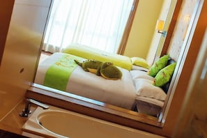 A hotel room with a bed, green pillows, a green blanket, and a mirror reflecting the room.