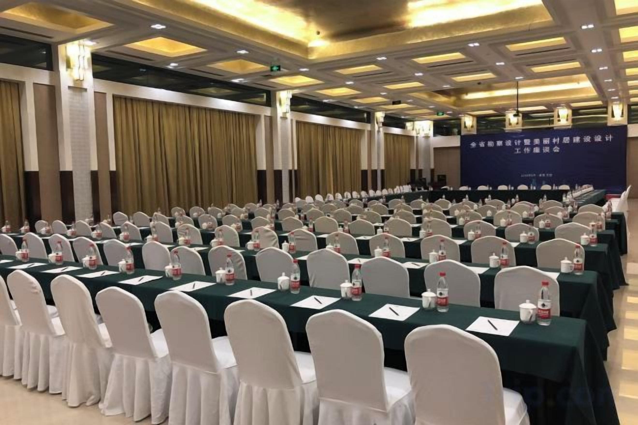 A conference room with rows of tables and chairs, a stage with a banner, and a ceiling with recessed lighting.
