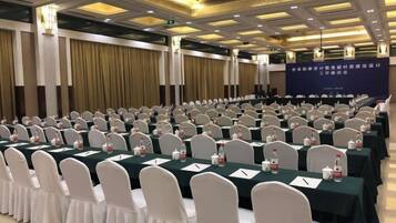 A conference room with rows of tables and chairs, a stage with a banner, and a ceiling with recessed lighting.