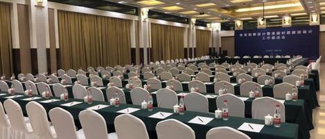 A conference room with rows of tables and chairs, a stage with a banner, and a ceiling with recessed lighting.