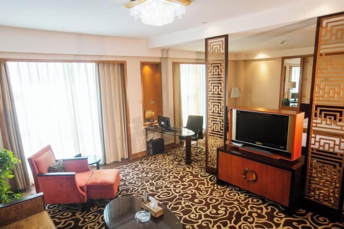 Une chambre d’hôtel équipée d’une grande télévision, d’un bureau et d’une chaise. Le sol est recouvert d’un tapis à motifs et un panneau décoratif orne le mur.