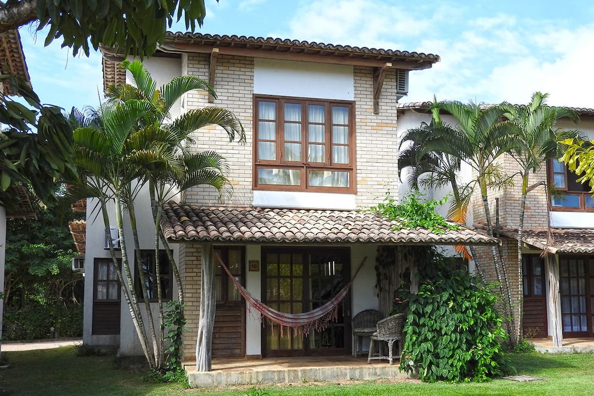 Una casa de dos pisos con techo de tejas, un porche con una hamaca y un pequeño jardín con palmeras.