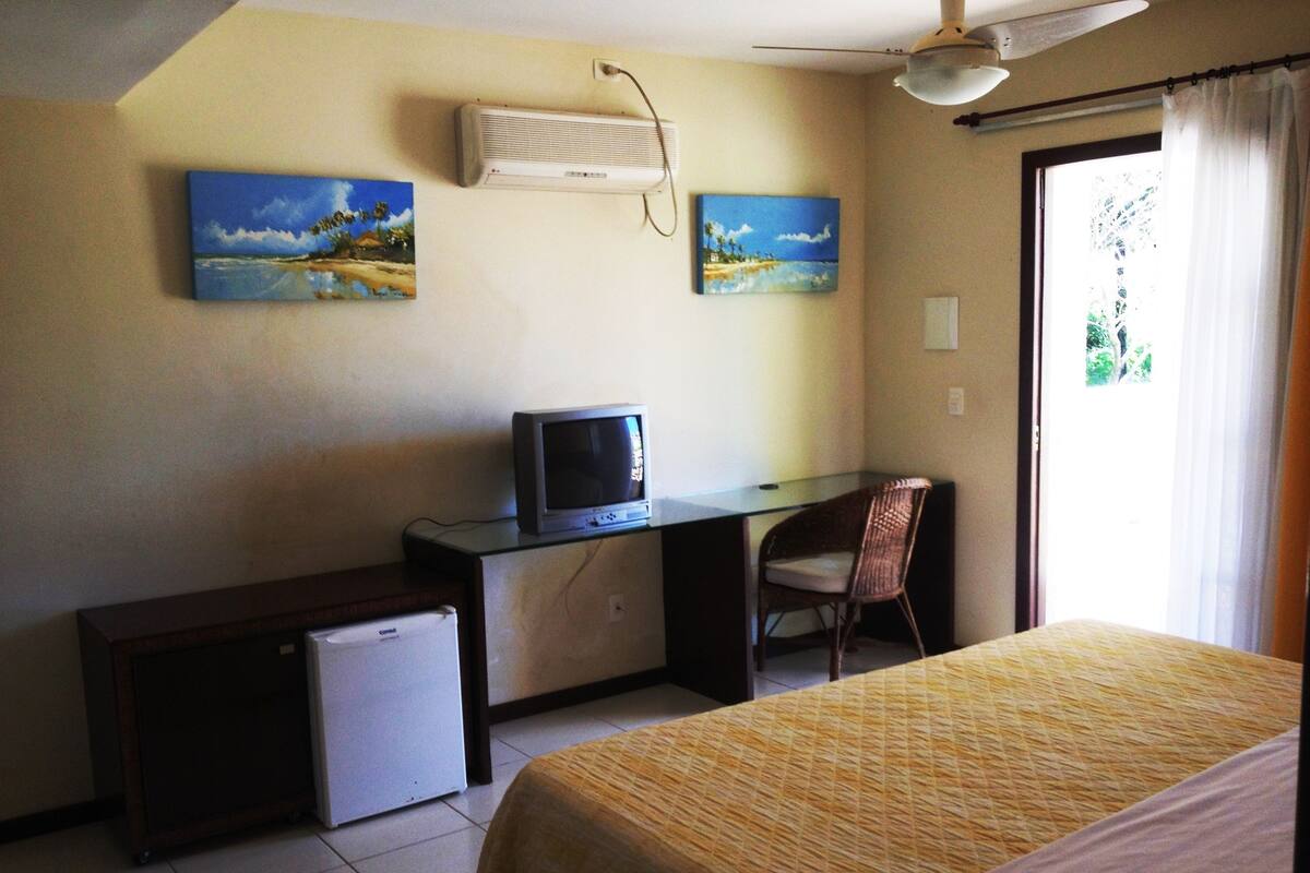 Una habitación de hotel con una cama, un escritorio con televisor, un refrigerador pequeño y dos cuadros en la pared.