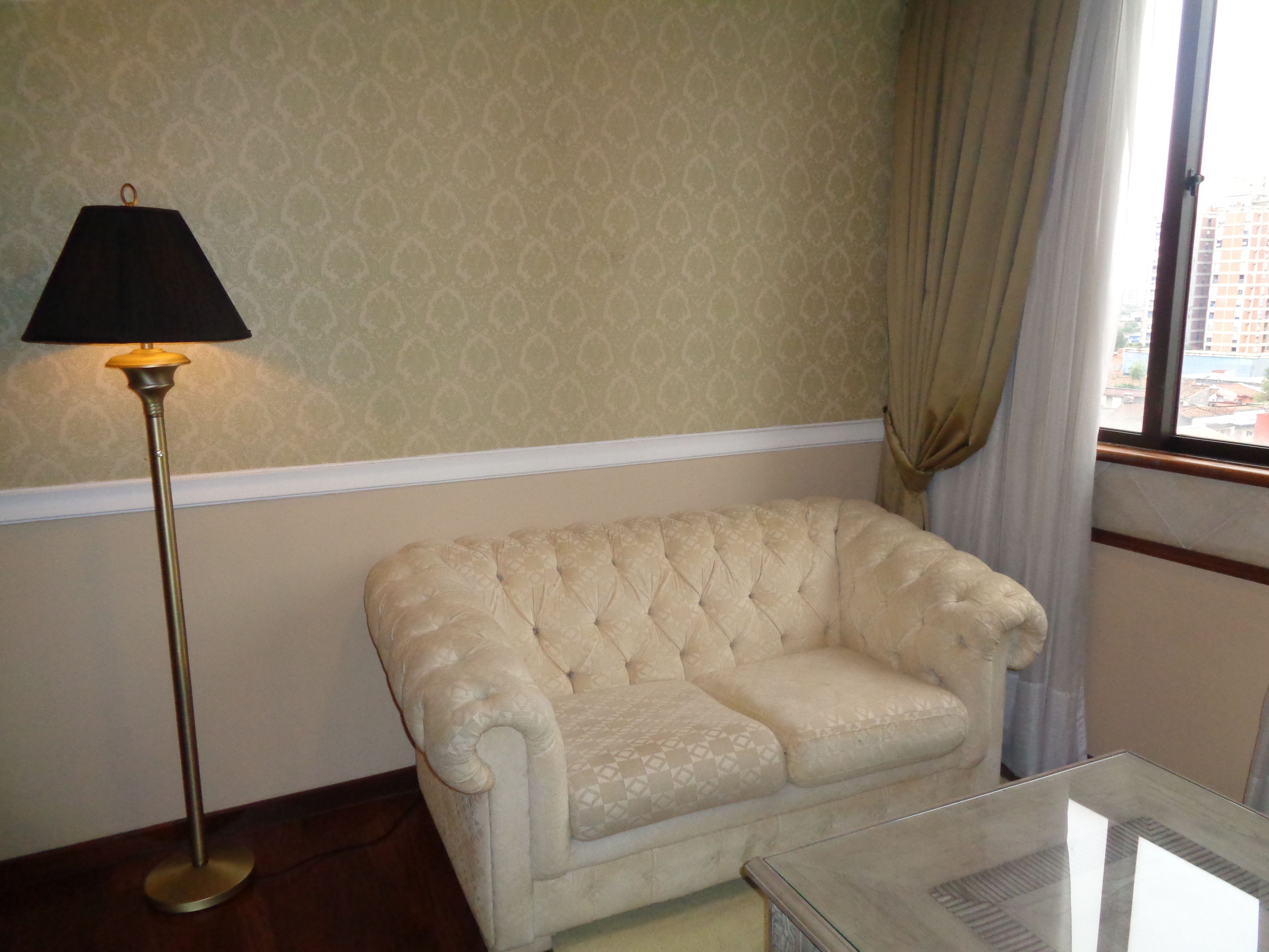 A room with a patterned wallpaper, a floor lamp, a tufted armchair, and a window with curtains.