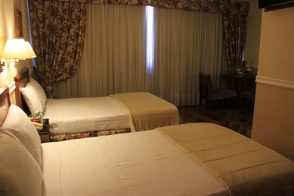Habitación de hotel con dos camas, una mesita de noche con lámpara, una silla y una pequeña mesa con fruta.