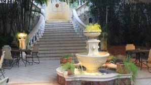 A fountain with a stone base surrounded by potted plants, a staircase leading up to a building, and outdoor seating with chairs and tables. - Nobile Hotel Premier Hill (Asunción)