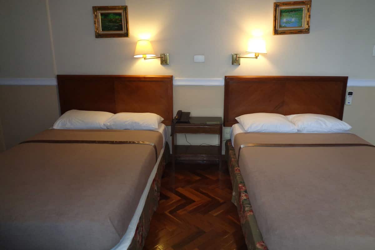A hotel room with two single beds, wooden headboards, bedside tables with lamps, and two framed pictures on the wall.