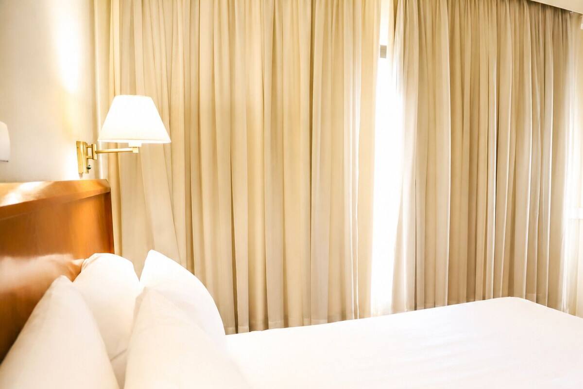 A hotel room with a bed, pillows, a lamp, and curtains.