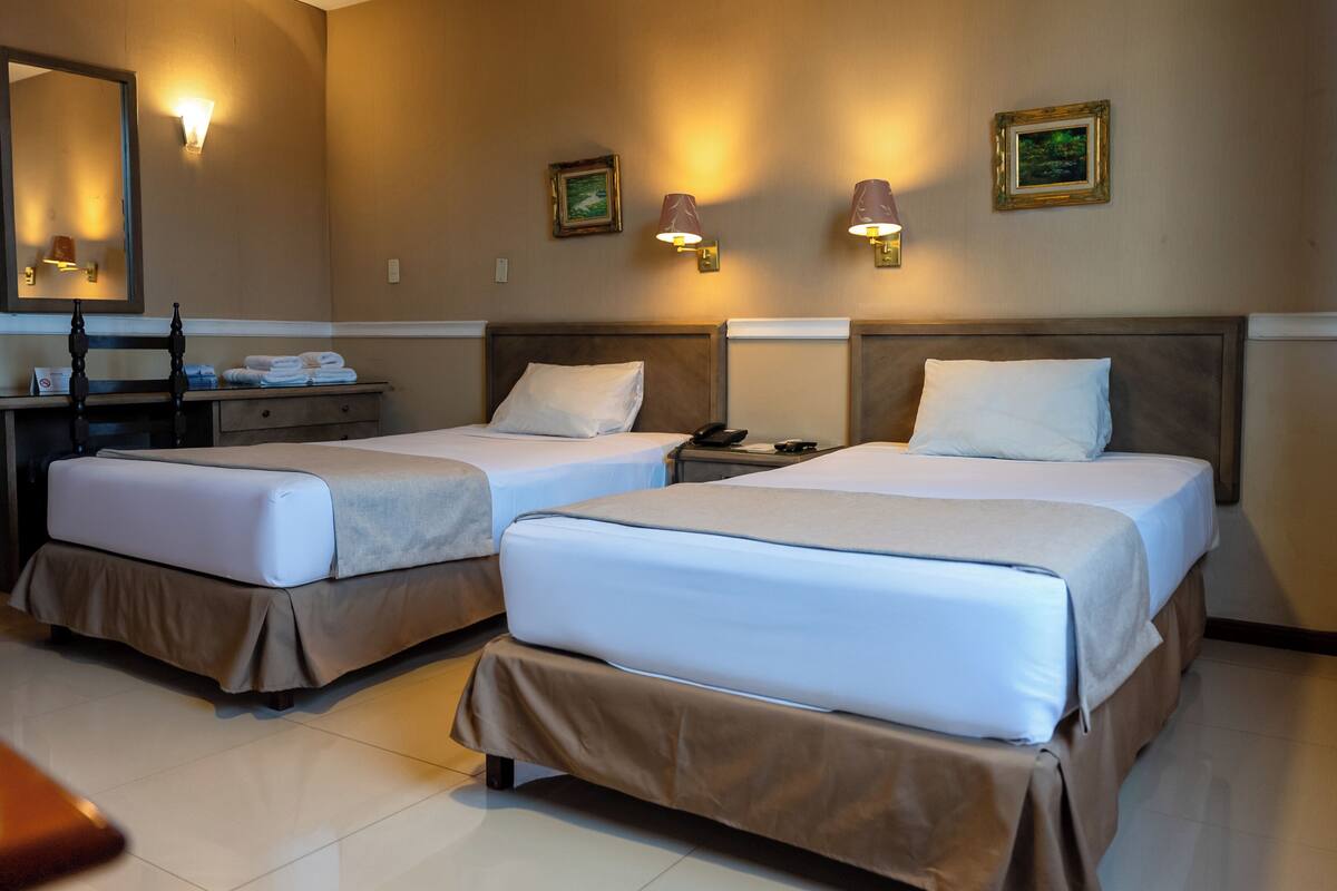 A hotel room with two beds, a desk, a mirror, and wall-mounted lamps.