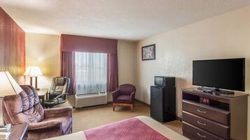 Une chambre d’hôtel comprenant un lit, deux chaises, un bureau avec une lampe, un téléviseur et une horloge numérique.