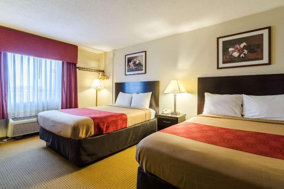 A hotel room with two beds, a window with curtains, a wall-mounted painting, and a bedside table with a lamp.