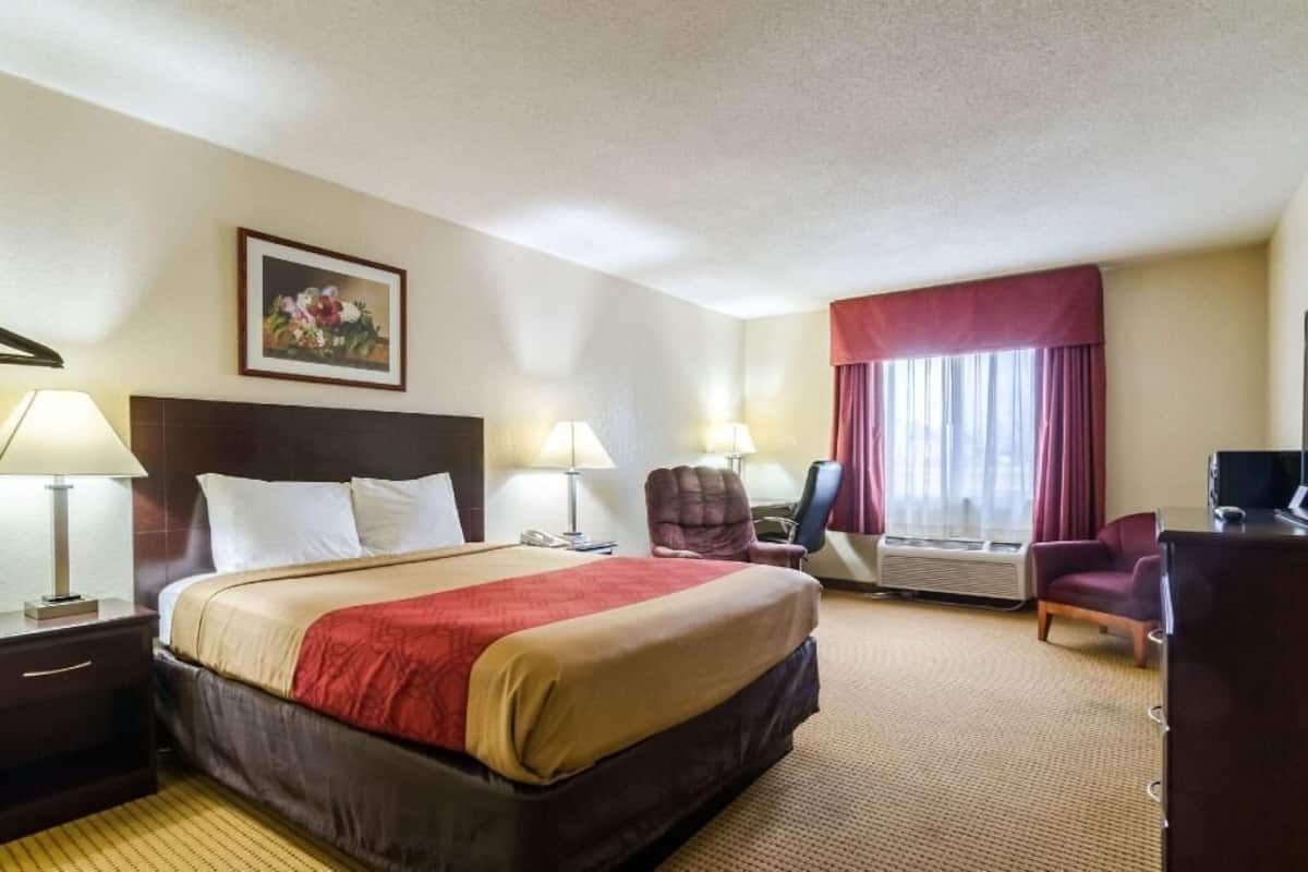 A hotel room with a large bed, two bedside tables with lamps, a desk with a chair, a television, and a window with curtains.