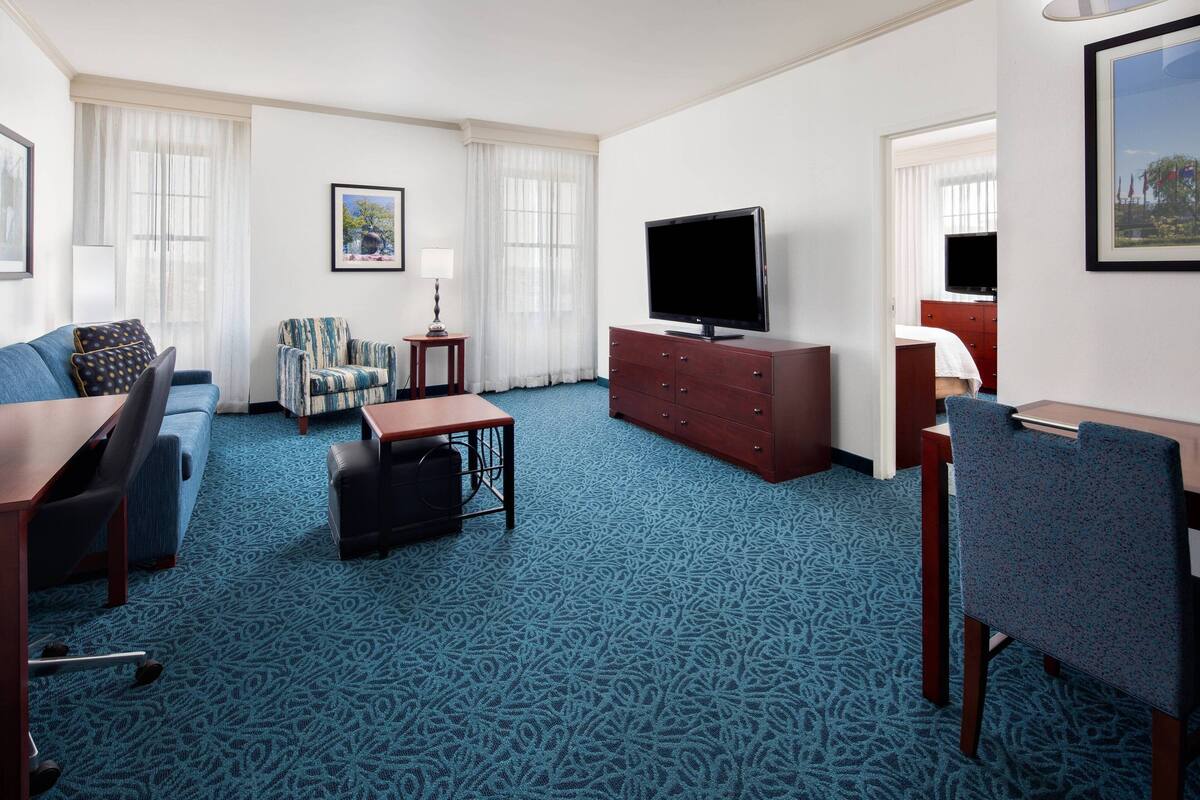 A hotel room with a blue patterned carpet, a sofa, a chair, a small table, a TV on a wooden cabinet, and a framed picture on the wall.
