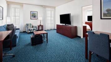 A hotel room with a blue patterned carpet, a sofa, a chair, a small table, a TV on a wooden cabinet, and a framed picture on the wall.