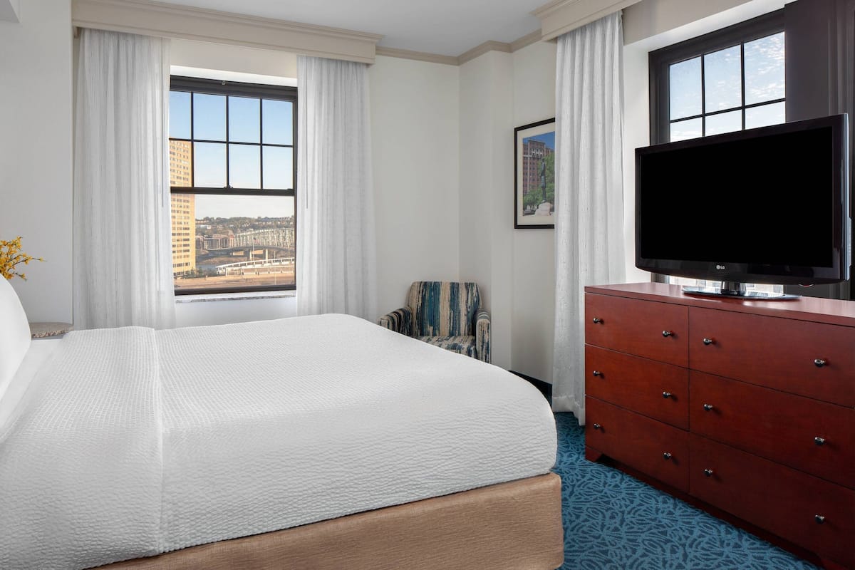 A hotel room with a large bed, a TV on a wooden dresser, and a window with a city view.
