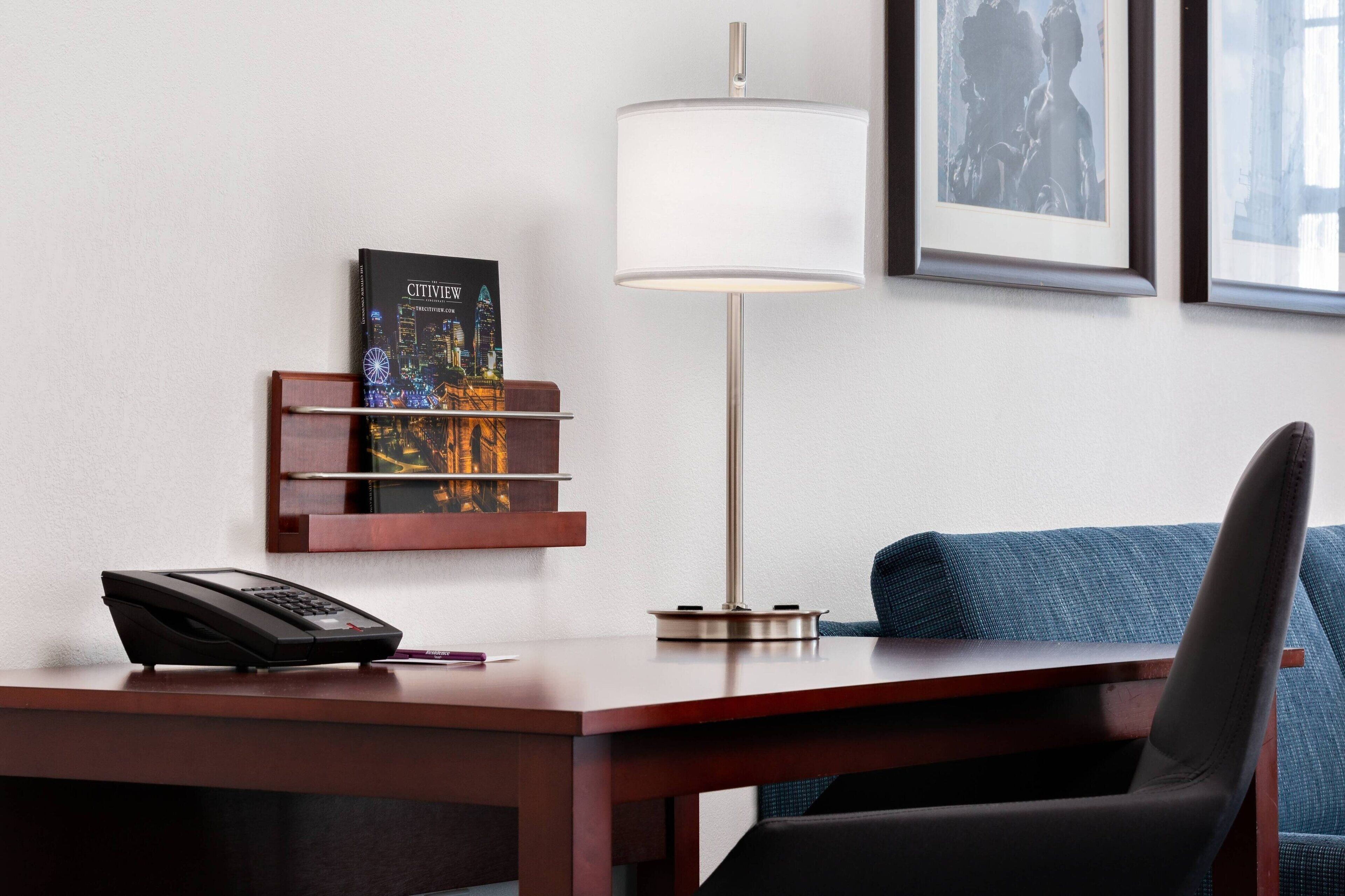 A hotel room with a desk, a lamp, a phone, a chair, a sofa, a bookshelf with a book, and framed pictures on the wall.