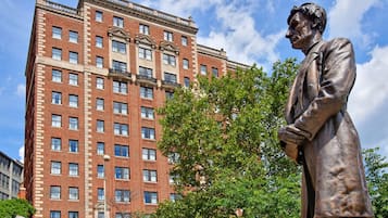 A statue of a man in a suit standing in front of a tall brick building with multiple windows and balconies.