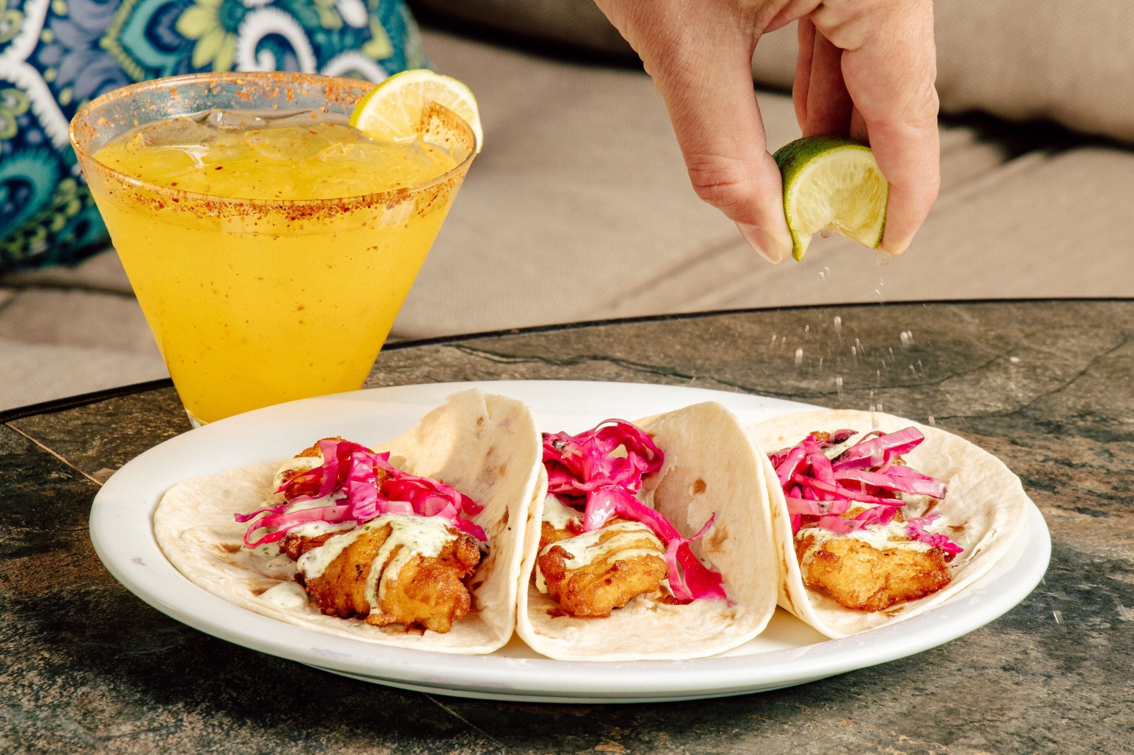 A plate of tacos with a lime being squeezed over them, and a glass of orange beverage with a slice of lime on the rim.