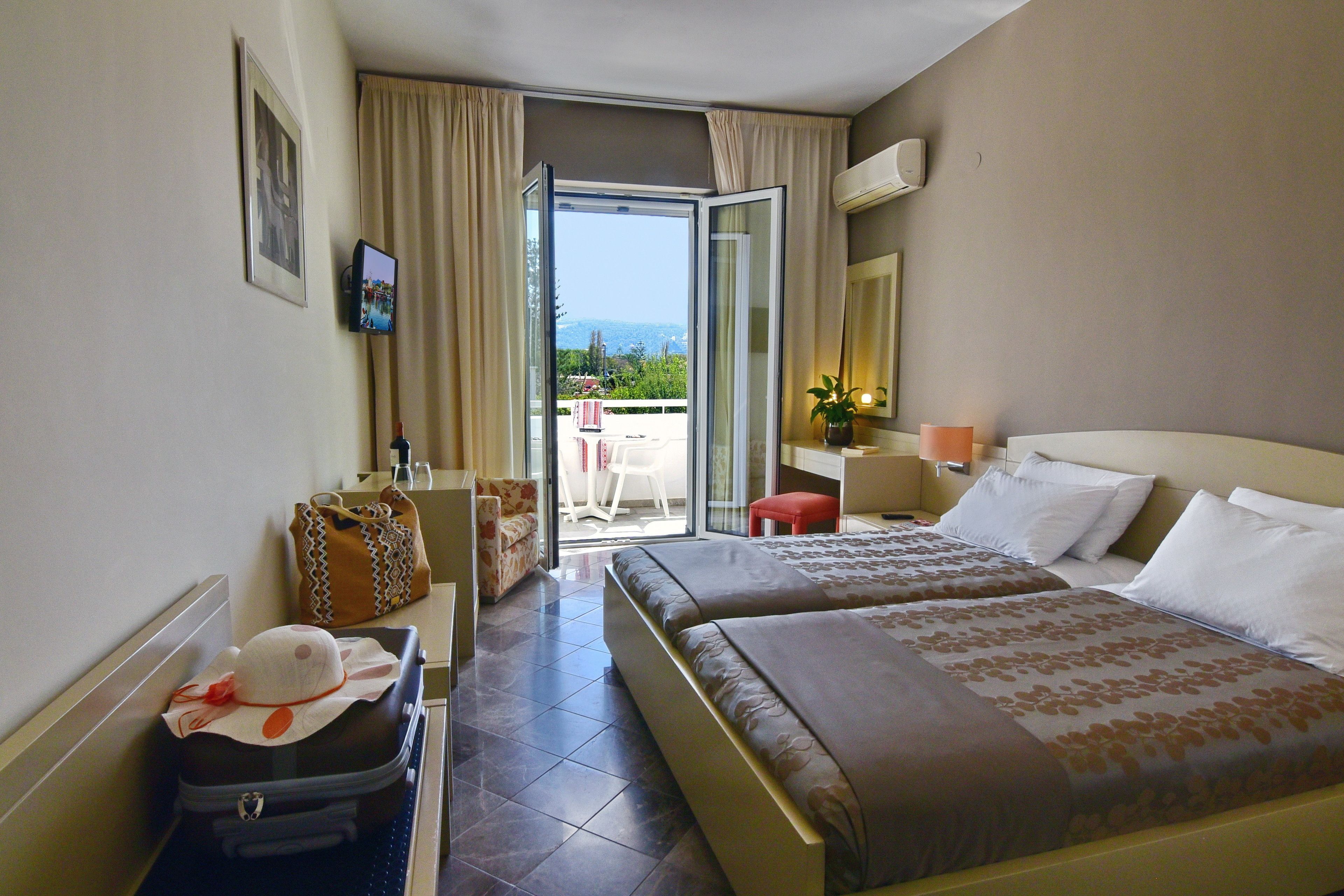 Habitación de hotel con dos camas, un escritorio, una silla, una maleta y un balcón con vistas.